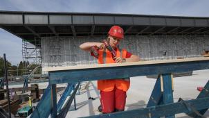 <p>Eine Frau schraubt bei einer Vorbesichtigung der Baustelle an einem Bauzaun.</p>