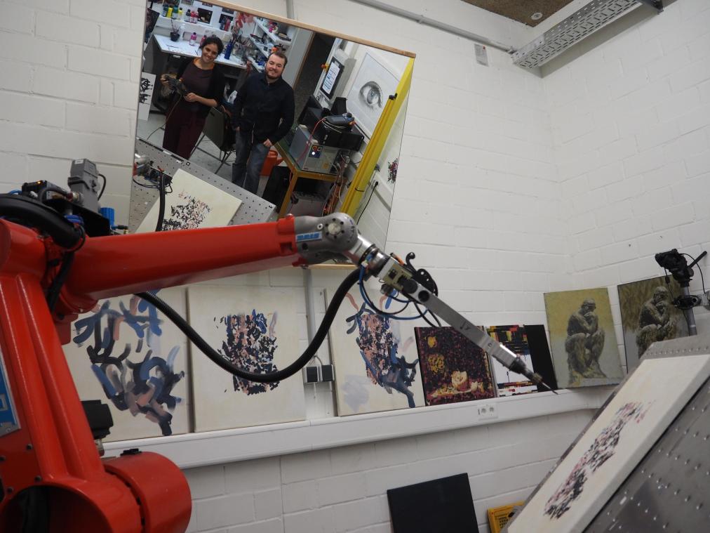 <p>Zu Anschauungszwecken befindet sich der Roboter e-David der Universität Konstanz in einer Mal-Position über der Leinwand, während der Computerwissenschaftler Marvin Gülzow und die Künstlerin Liat Grayver über einen Spiegel in die Kamera blicken.</p>