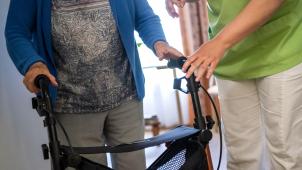 <p>Eine Pflegefachkraft hilft einer Bewohnerin in einem Seniorenheim mit ihrem Rollator.</p>