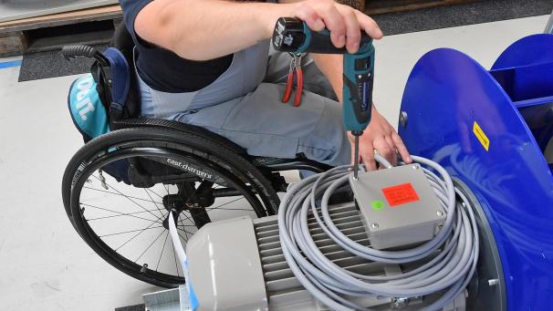 <p>An einem Arbeitsplatz für Motorklemmen ist ein Mitarbeiter im Rollstuhl mit Montagearbeiten beschäftigt.</p>