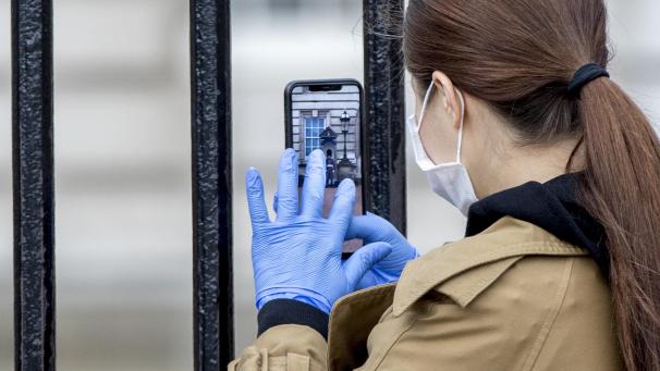<p>Eine Besucherin trägt Handschuhe und Mundschutz, während sie durch die Gitterstäbe vor dem Buckingham Palast einen Queens Guard fotografiert.</p>