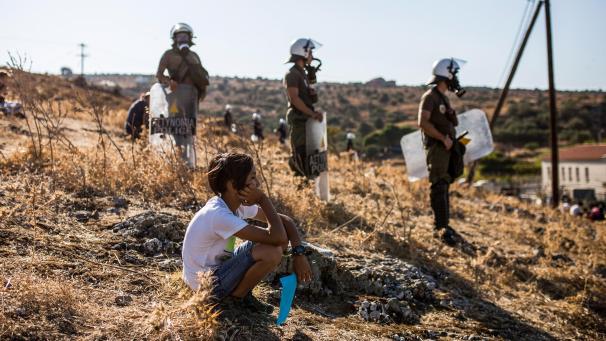<p>Ein Junge sitzt vor Polizisten und beobachtet einen Protest von Migranten nach den Nachrichten über die Schaffung eines neuen provisorischen Flüchtlingslagers auf der Insel Lesbos hoch.</p>