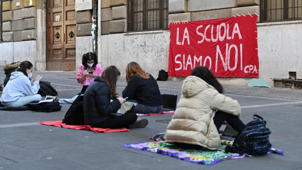 <p>Schülerinnen sitzen vor ihrem Gymnasium, an dem ein Banner mit der Aufschrift „La Scuola siamo Noi“ (Zu Deutsch: „Die Schule sind wir“) hängt.</p>