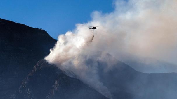 <p>Ein Militärhubschrauber wirft Wasser auf einen brennen Berggipfel ab.</p>