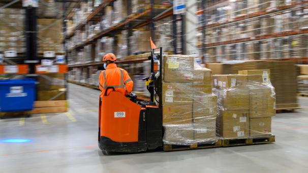 <p>Ein Mitarbeiter des Amazon Logistikzentrums fährt mit Paketen an Hochregalen vorbei.</p>