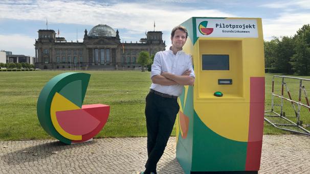 <p>Michael Bohmeyer, Initiator des Vereins „Mein Grundeinkommen“, steht an einem symbolischen Geldautomaten, im Hintergrund das Reichstagsgebäude.</p>