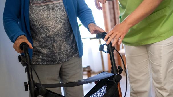 <p>Eine Pflegefachkraft hilft einer Bewohnerin in einem Seniorenheim mit ihrem Rollator.</p>