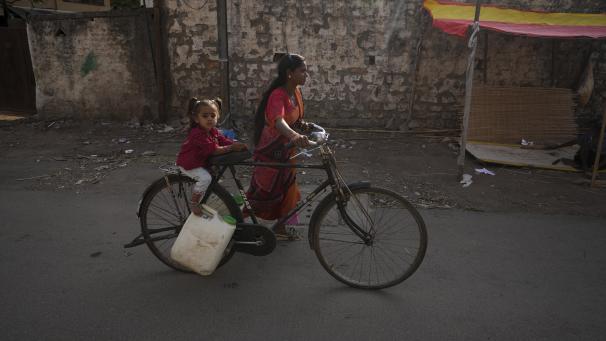 <p>Eine Frau mit einem Kind transportiert Wasser mit einem Fahrrad aus einem öffentlichen Wasserhahn in einem indischenSlumgebiet.</p>