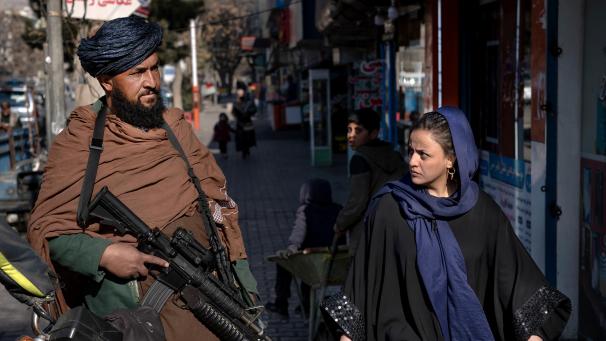 <p>Ein Taliban-Kämpfer steht Wache während eine Frau vorbeiläuft.</p>