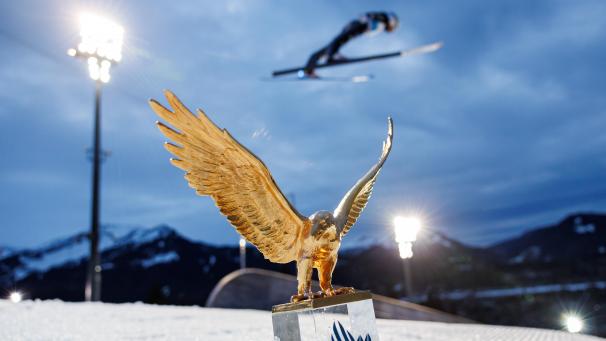 <p>Der goldene Adler, der Pokal für den Gesamtsieger der Vierschanzentournee, steht im Schnee vor einem Athleten in Aktion.</p>