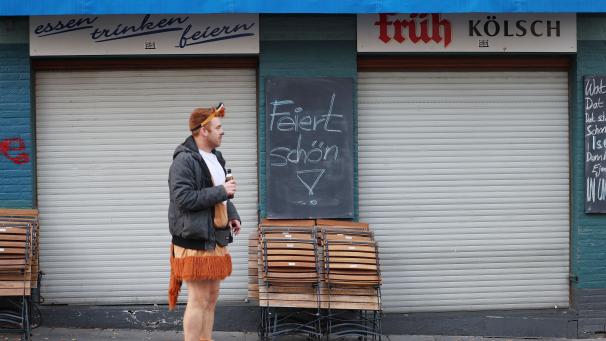 <p>Ein Karnevalist steht vor einer geschlossenen Kneipe, an der ein Schild mit der Aufschrift „Feiert schön!“ hängt.</p>