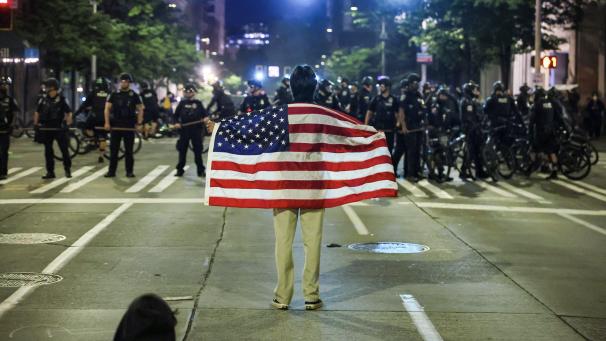 <p>Ein einsamer Demonstrant mit einer amerikanischen Flagge steht vor einer Polizeikette, nachdem er in der Innenstadt von Seattle den Befehl erhalten hat, sich zu entfernen.</p>