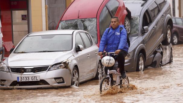 <p>Ein Mann radelt im Oktober 2024in Valencia durch überflutete Straßen an Autos vorbei, die von den Wassermassen übereinander geschoben wurden.</p>