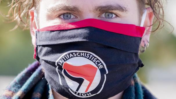 <p>Ein Mann mit Antifa-Maske bei einer Demo in Potsdam. Das Logo der Antifa-Bewegung zeigt zwei nach links gerichtete, gegen rechts geneigte Fahnen: eine rote, die für den Sozialismus/Kommunismus steht und eine schwarze, die den Anarchismus symbolisiert.</p>
